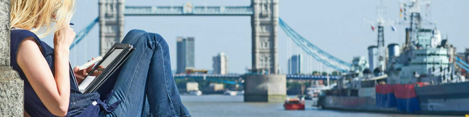 A blonde student wearing a navy tank top and jeans sits on a stone wall in her bare feet reading on a tablet computer in front of the iconic Tower Bridge in London.